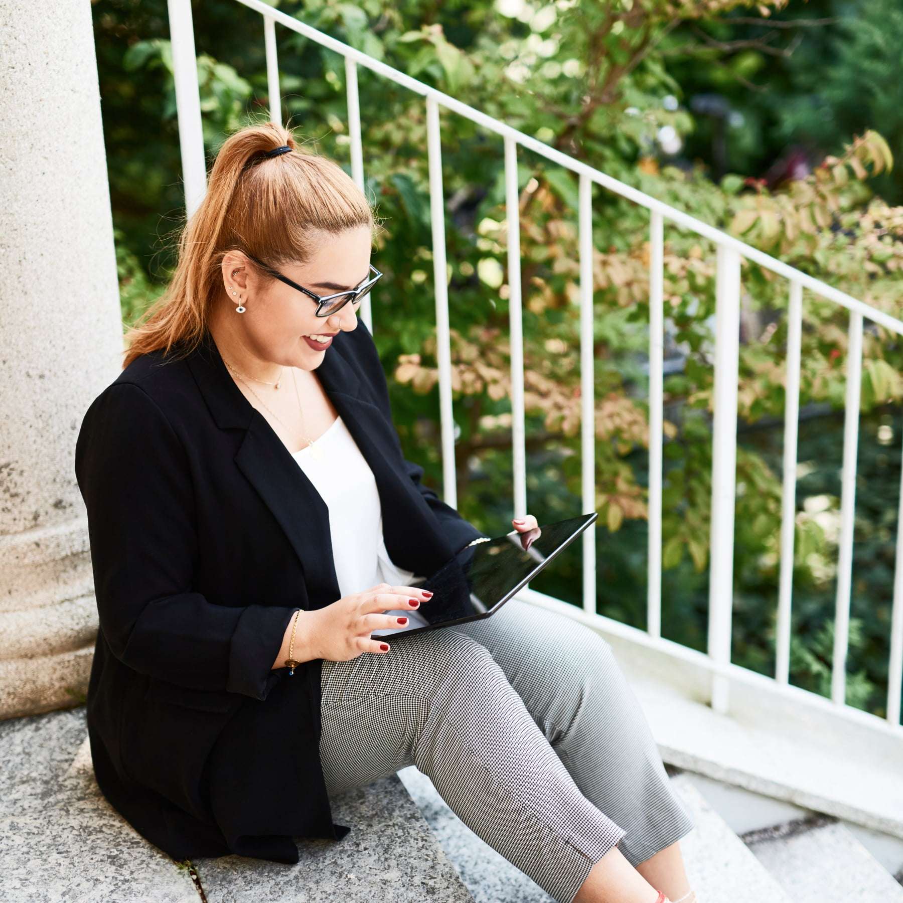 A woman sits on a staircase with white railings in front of trees and holds a tablet in her hand, which she operates.
