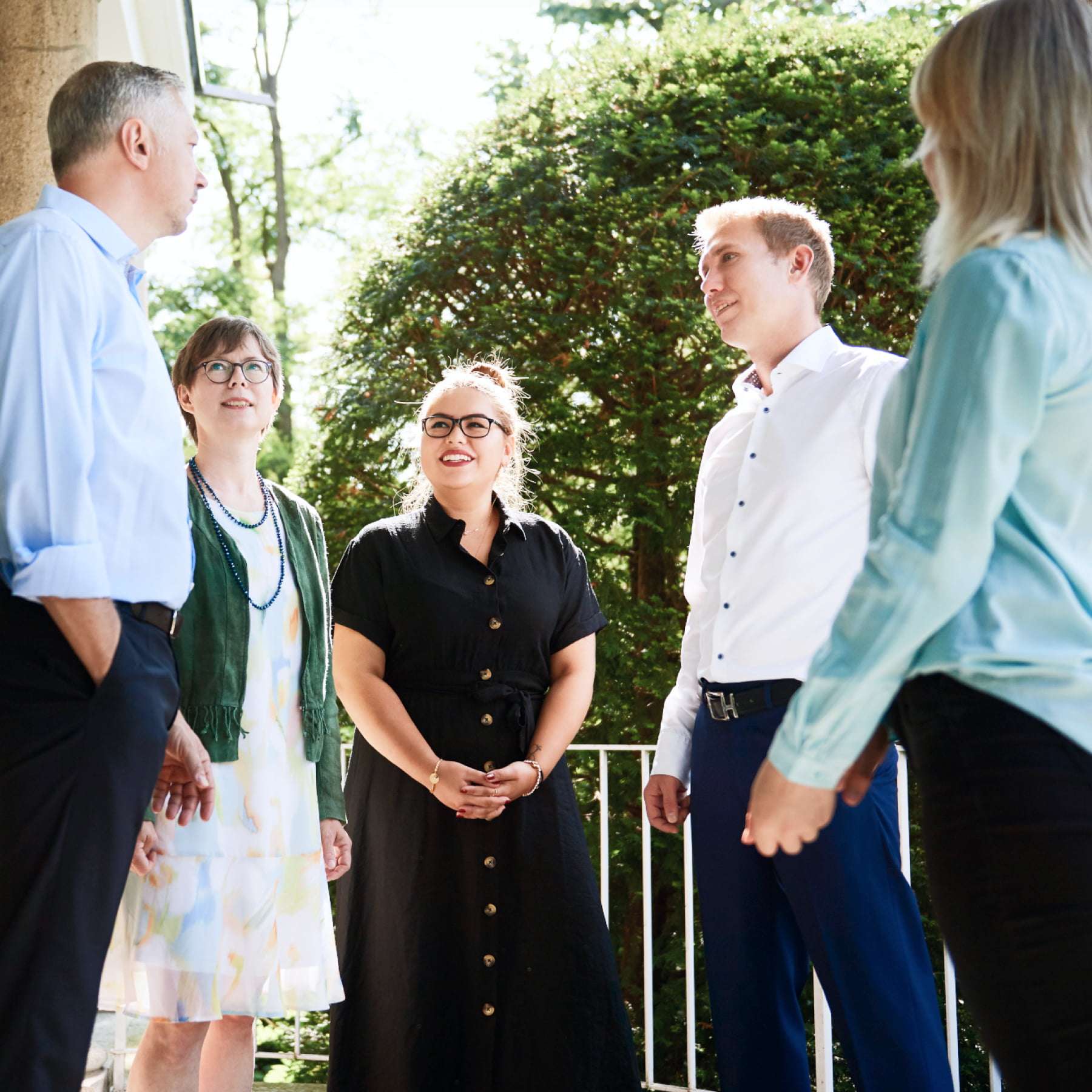 2 men and 3 women are standing on a balcony in front of green trees and talking. The sun is shining.
