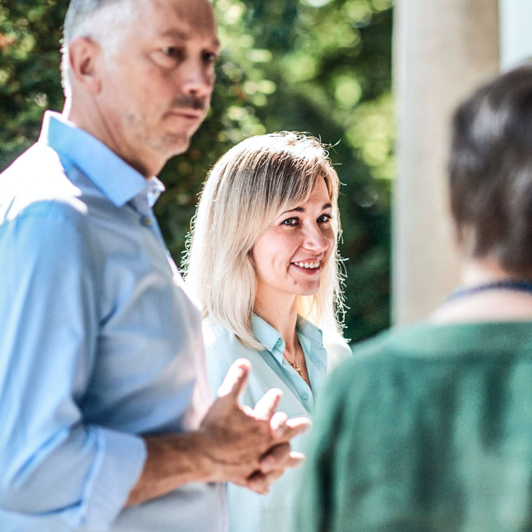 A man and two women are standing next to a pillar and in front of green trees, engrossed in conversation