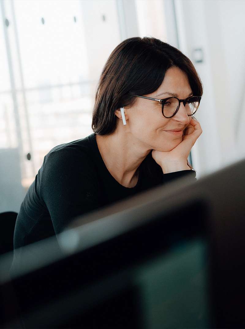 Annika is smiling as she works at her PC, resting her chin on her left hand. She can be seen from the side and is wearing an earpod in her right ear.