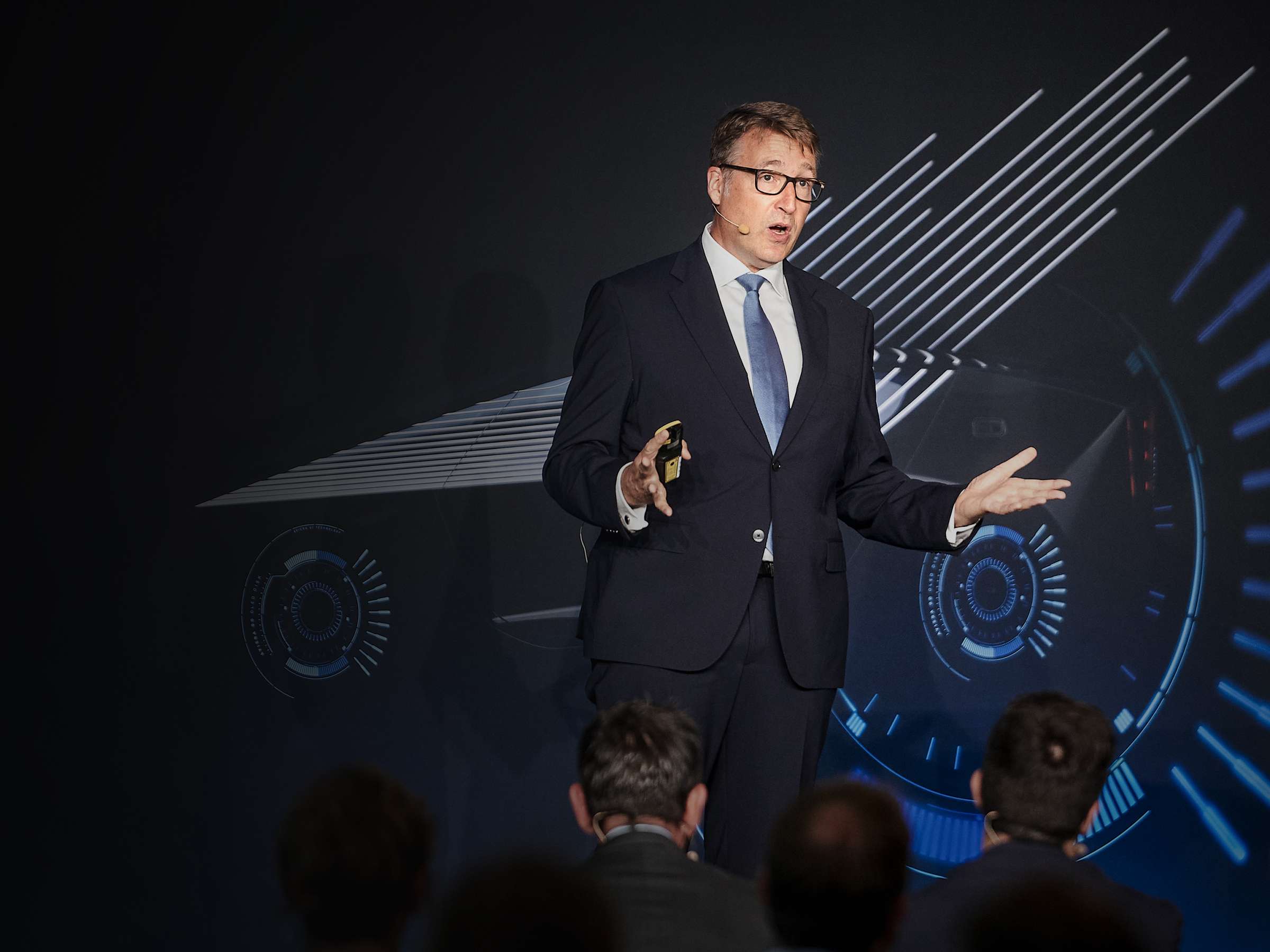 A man in a suit stands on a stage and gives a lecture to an audience that is only vaugly perceptible, gesticulating with both hands.