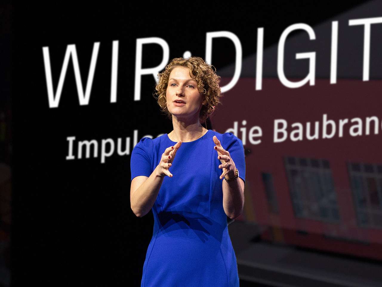 A woman in a blue dress giving a talk at a trade fair on impulses for the construction industry and gesturing with her hands.