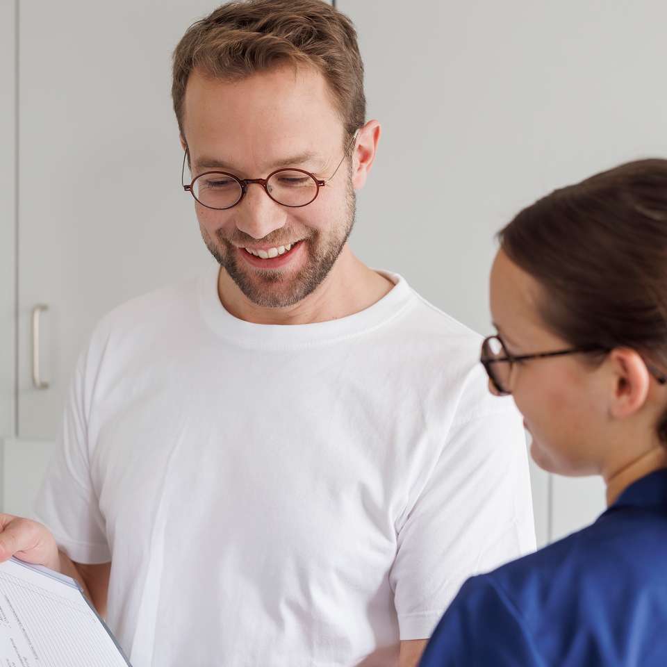 A man wearing glasses and a white t-shirt is smiling while talking to a woman in blue clothing and looking at a document.