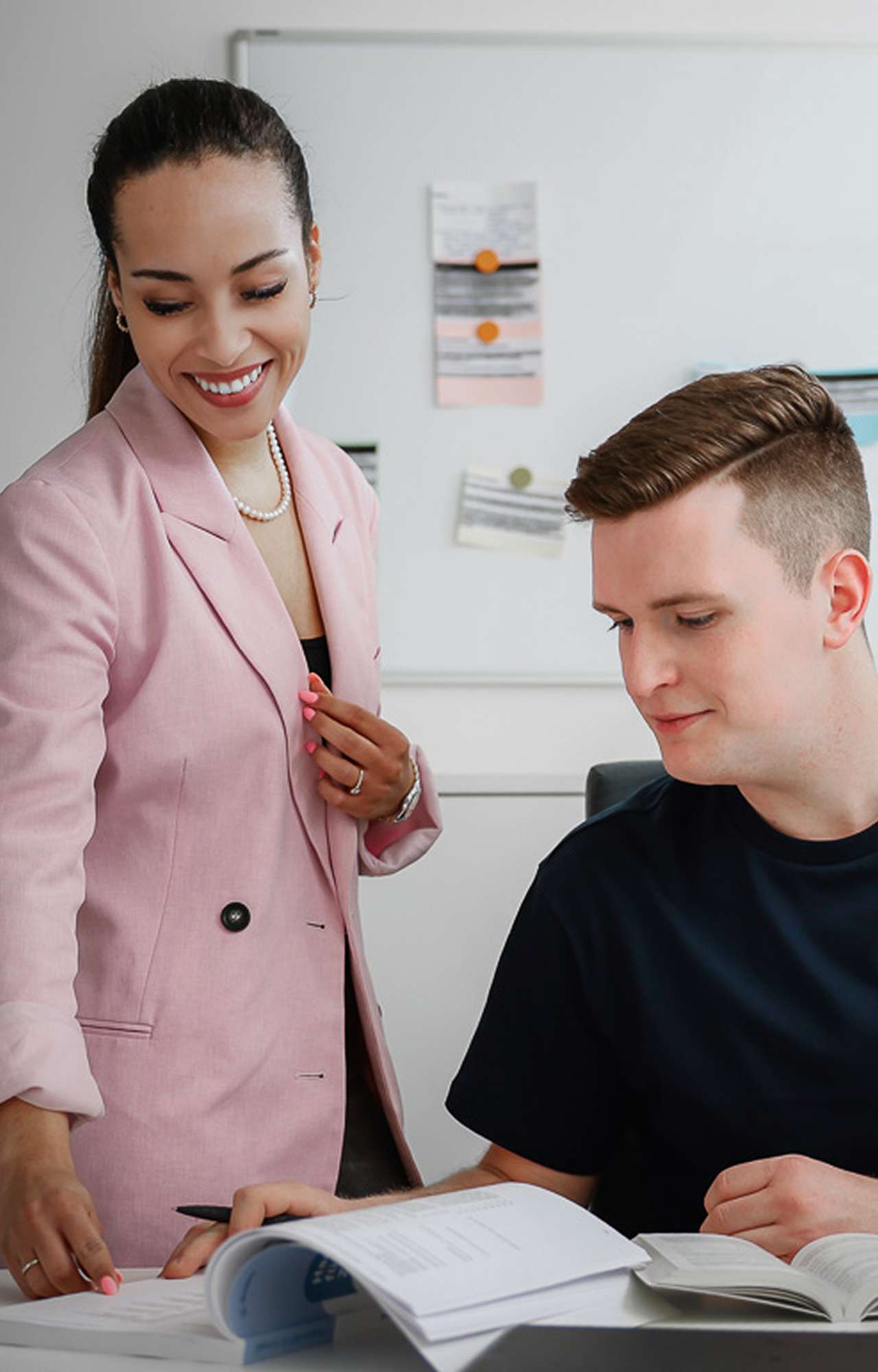 A woman in a pink blazer is standing next to a man who is working at a table with documents.