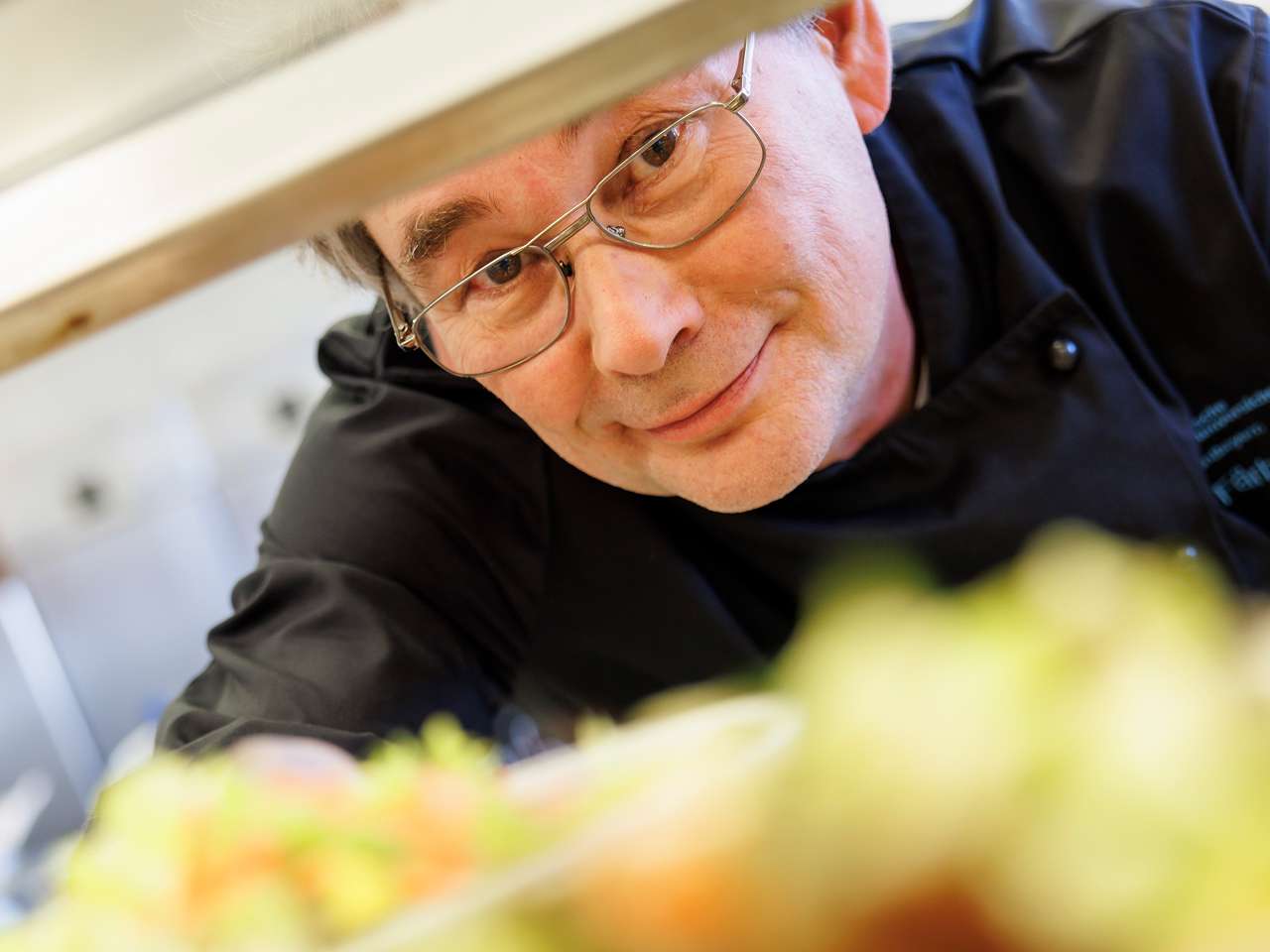 Chef in a kitchen, slightly leaning against a counter and smiling at the camera.