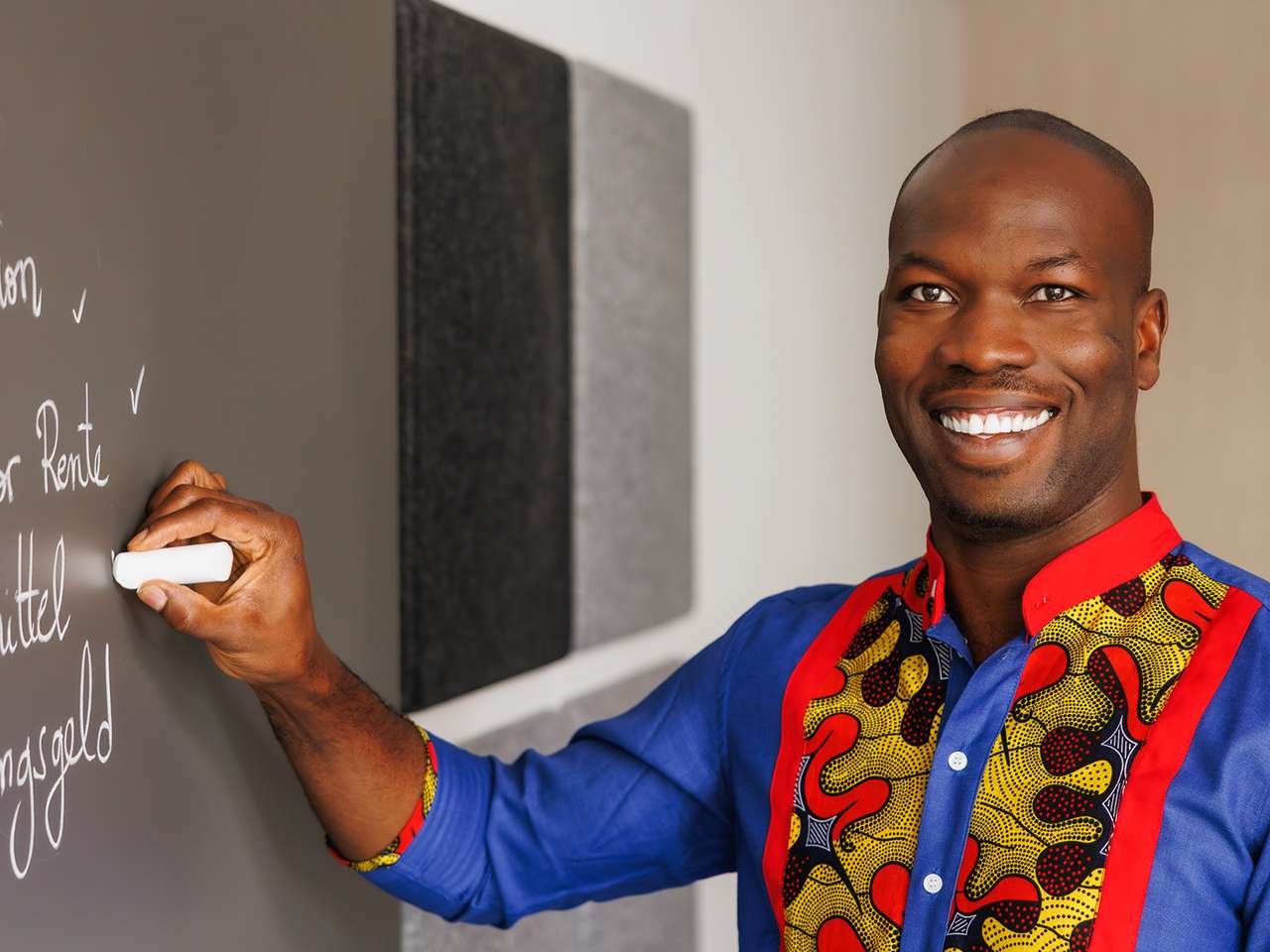 Smiling man in a colourful shirt writing on a chalkboard.