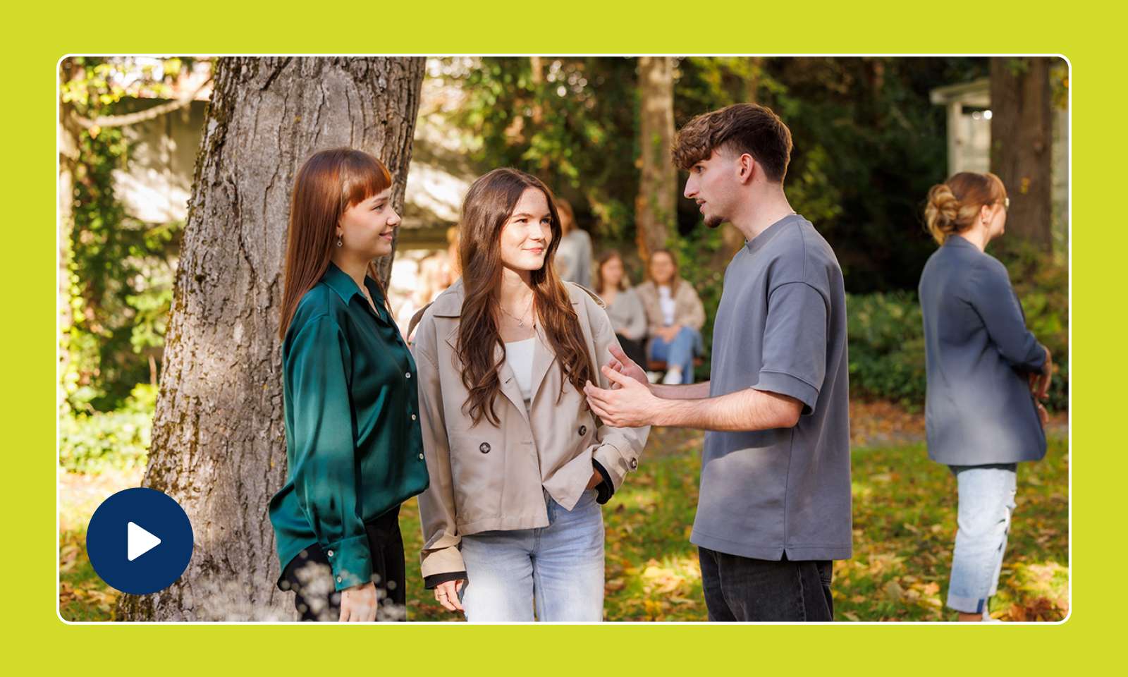 Three young adults are standing under a tree and chatting with smiles on their faces, while more people can be seen in the background.