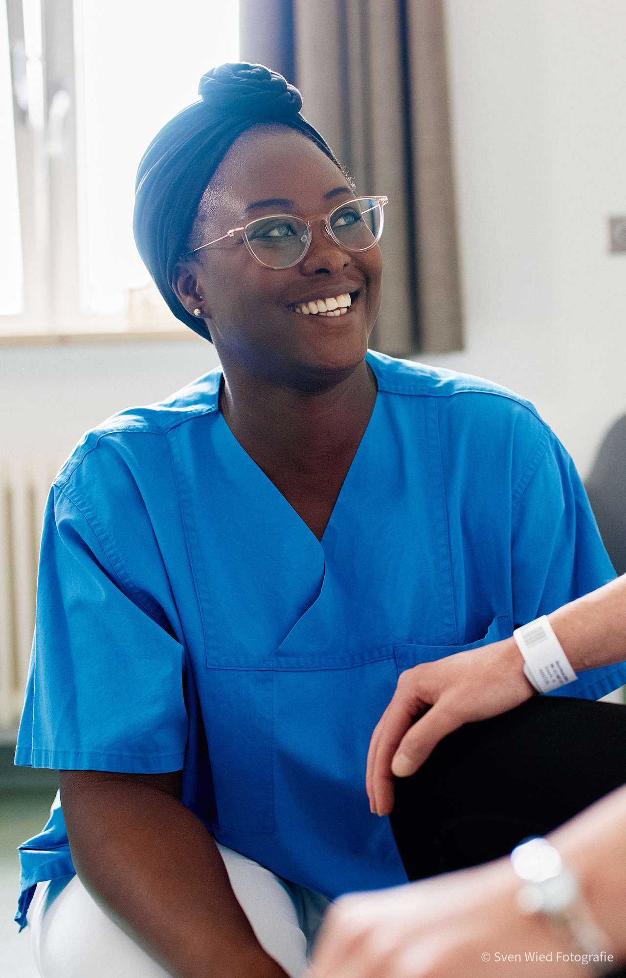 Smiling person wearing blue clothing and headwear, talking to another person in a bright room. 