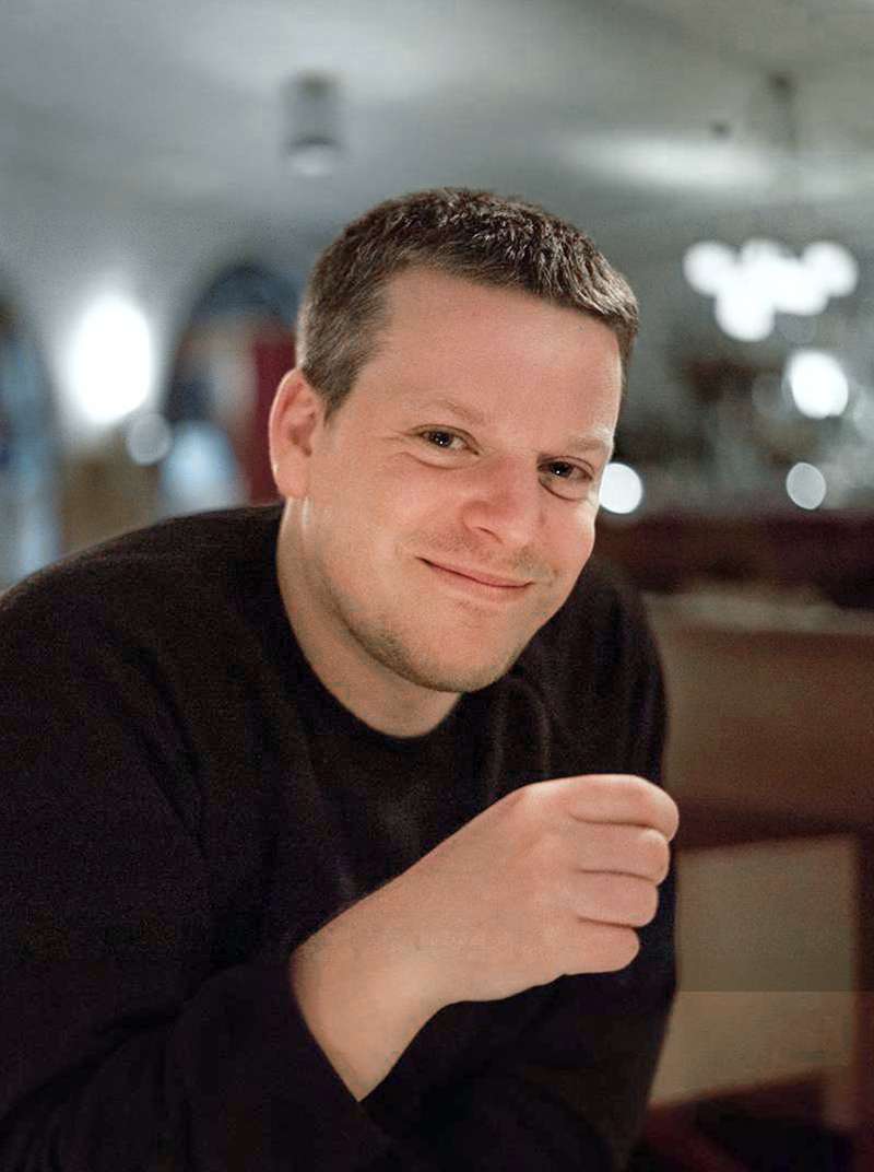 Markus sits smiling in a restaurant with his right hand raised in front of his upper body.
