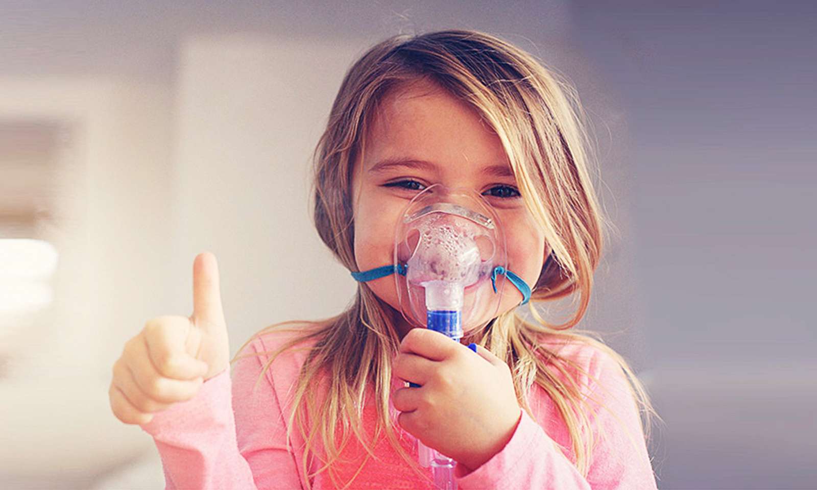 Approx. 4-year-old child with blue inhalation mask sticks his thumb up and laughs under the mask
