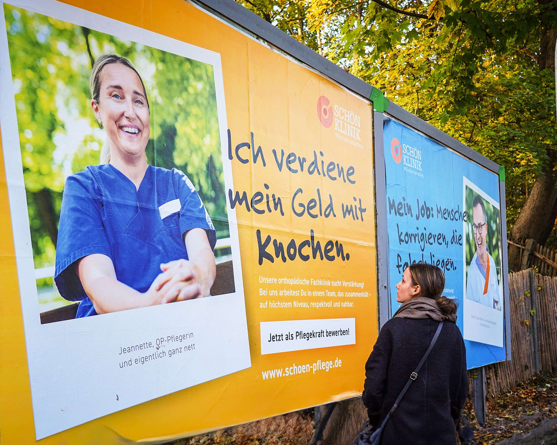 Eine Frau schaut sich interessiert zwei nebeneinander hängende Straßenplakate (in orange/gelb und himmelblau) aus der Personalkampagne der Schön Klinik an